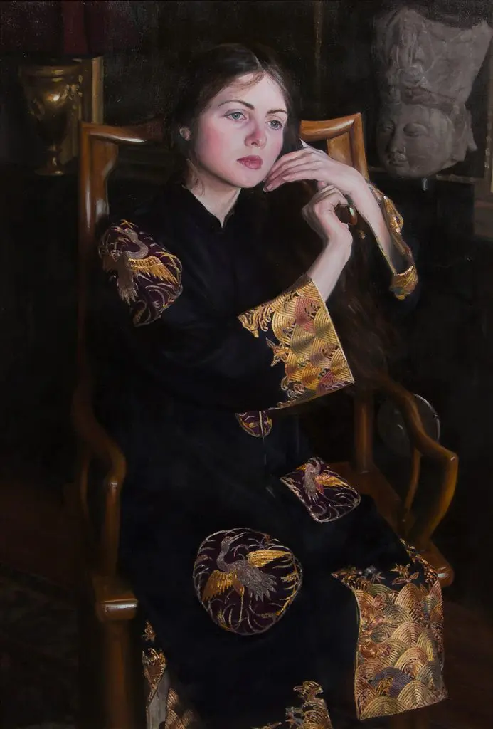 Woman wearing black gown with gold accents, sitting in a wooden chair in a darkly lit room, running her hands through her hair