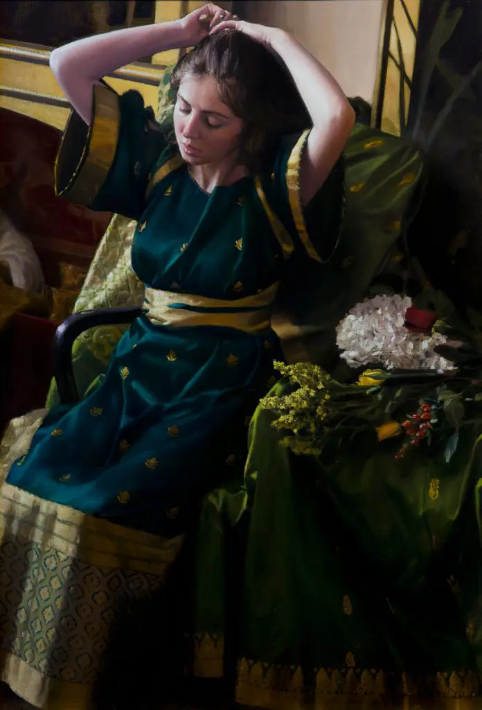 Woman coming her brown hair, wearing green gown with gold accents, sitting on a chair with a green tapestry hanging over it