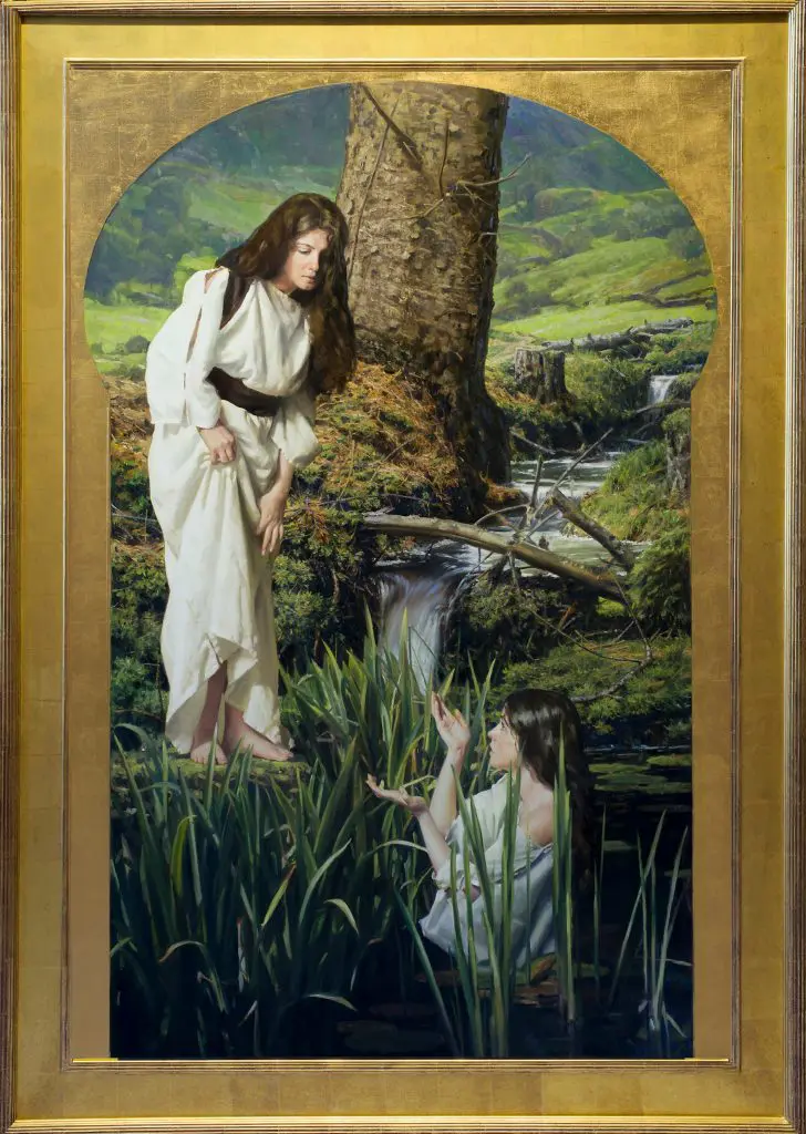 Brunette woman wearing a white robe, standing along the side of a stream, looking down at another brunette woman in white standing in the water