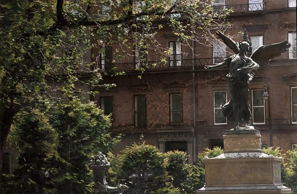 View of a city park with greenery and a statue of an angel in front of a red brick residential building