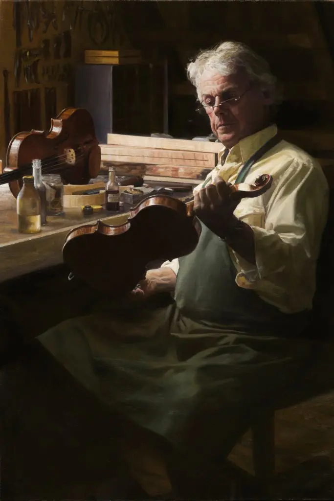 Man with white hair and glasses, wearing an apron, working on violin in wood shop