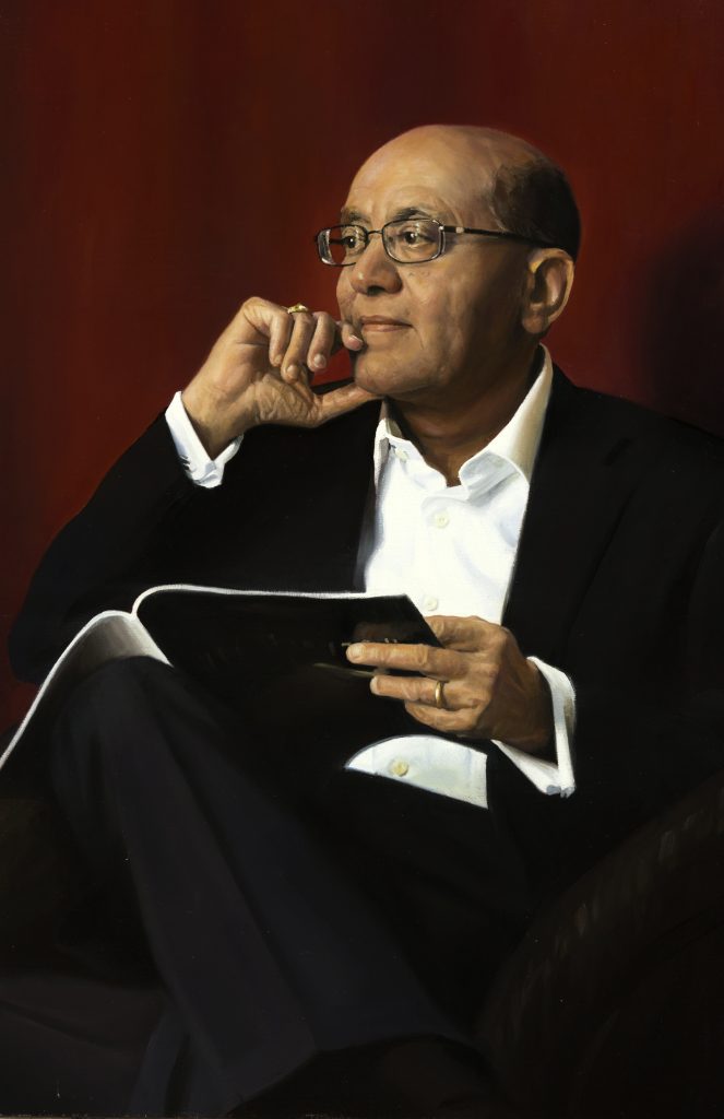 Dr. Rakesh Jain holding a notebook, sitting inquisitively in front of a red backdrop